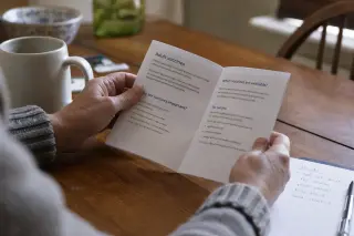 Hands of a middle-aged person reading a pharmacy leaflet about vaccines at a kitchen table in soft natural morning light, with a mug and to-do list nearby.