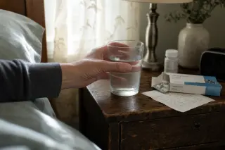 Adult's hand reaching for a glass of water on a wooden nightstand with medicine box and handwritten note in morning bedroom light