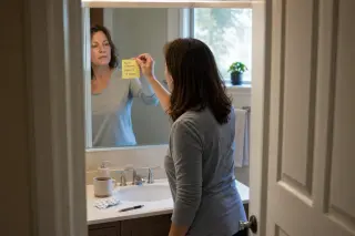 Middle-aged woman in a bathroom placing a screening reminder note on the mirror, early morning light, countertop with everyday items.