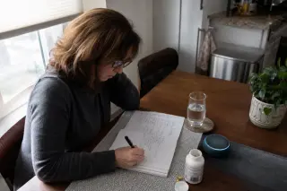 Older adult sitting at a kitchen table in morning light, hand turning pages of a diary marked with sticky notes, pill blister and pen nearby, warm natural atmosphere.